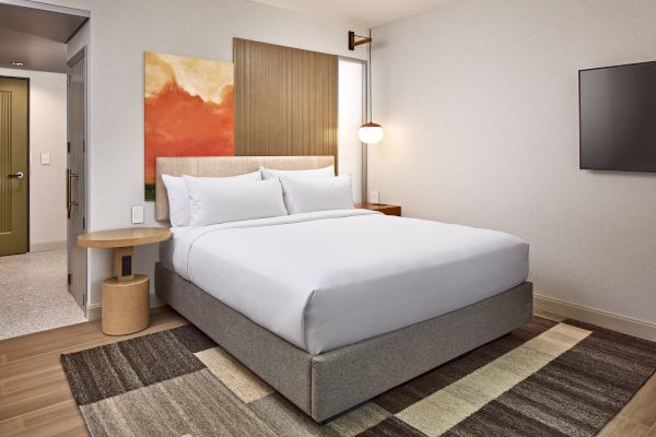 A modern hotel room with a neatly made bed, abstract artwork, wooden accents, a wall-mounted TV, and a stylish lamp.