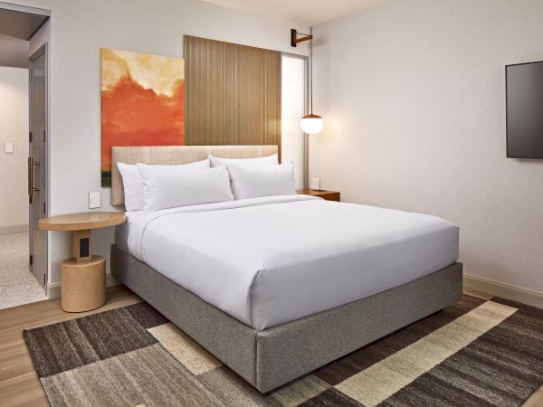 A modern hotel room with a neatly made bed, abstract artwork, wooden accents, a wall-mounted TV, and a stylish lamp.