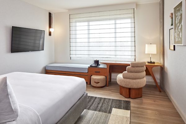 A modern hotel room with a bed, wall-mounted TV, window seat, desk, unique chair, lamp, and stylish decor.