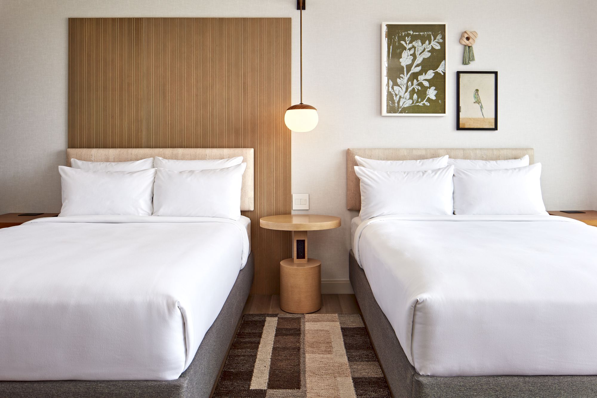 The image shows a modern hotel room with two neatly made beds, a central lamp, and artwork on the wall above the headboards.
