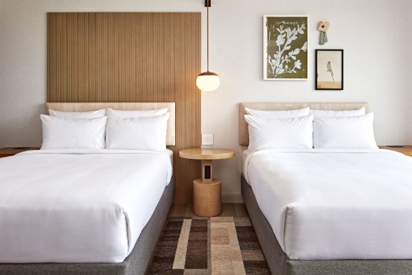 The image shows a hotel room with two neatly made beds, a wooden headboard, and decorative art on the wall with a hanging lamp above a table.