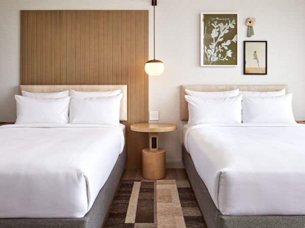 The image shows a hotel room with two neatly made beds, a wooden headboard, and decorative art on the wall with a hanging lamp above a table.
