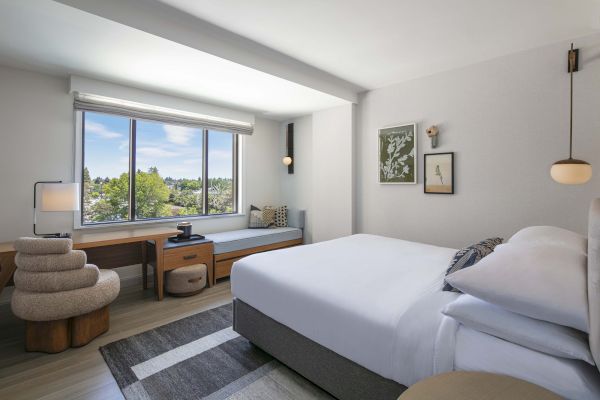 A modern bedroom features a bed, large window with a view, cozy corner seating, and a desk with a chair. Its decor is minimalist.