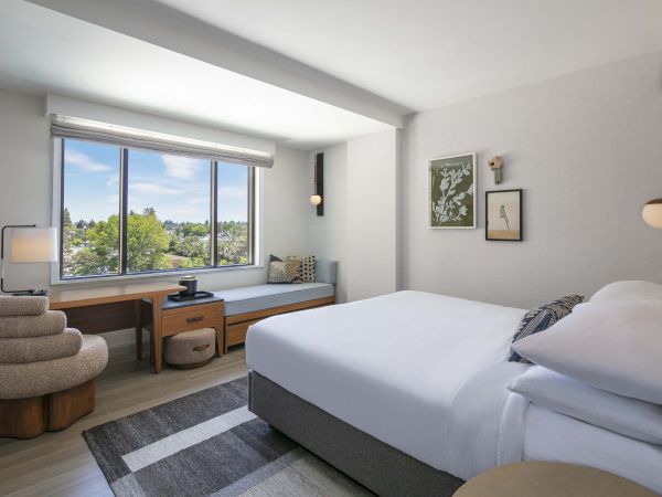 A modern bedroom features a bed, large window with a view, cozy corner seating, and a desk with a chair. Its decor is minimalist.