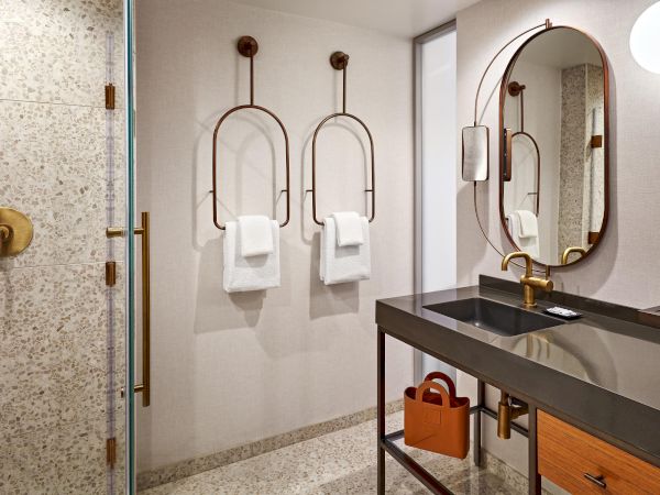 A modern bathroom with terrazzo tiles, sleek fixtures, a large oval mirror, towels on geometric racks, and a countertop with toiletries.