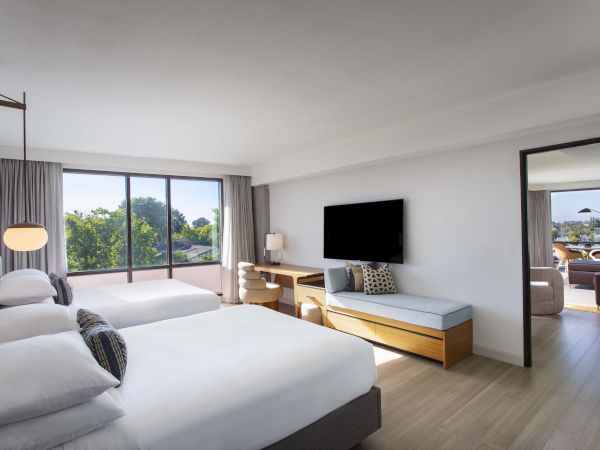 The image shows a hotel room with two beds, a TV, seating, and a view outside. The room leads to a seating area.