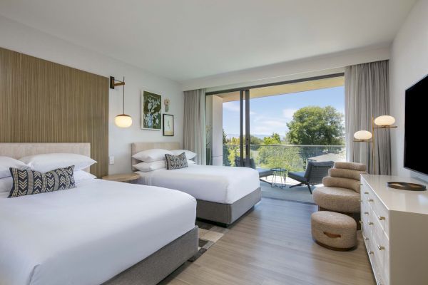 The image shows a hotel room with two beds, a balcony with a view, modern furniture, art on the walls, and a flat-screen TV.