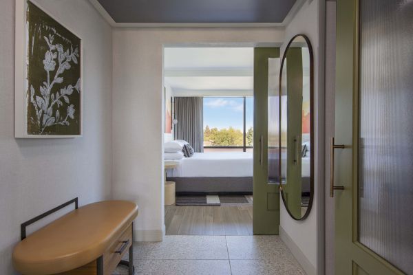 A hotel room hallway with artwork, a tan bench, and a mirror leading to a bedroom with a large window view.