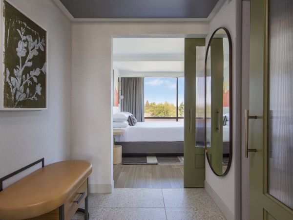 A hotel room hallway with artwork, a tan bench, and a mirror leading to a bedroom with a large window view.
