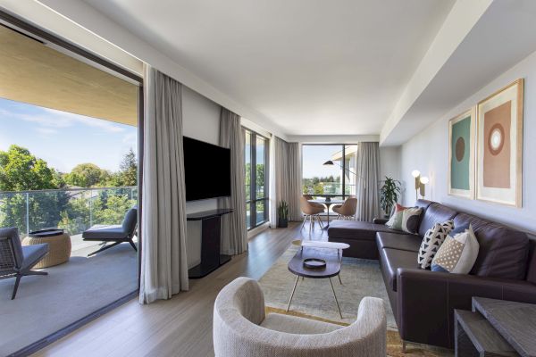 A modern living room with a balcony view, featuring a TV, sofas, armchair, abstract artwork, and a dining area with plants.