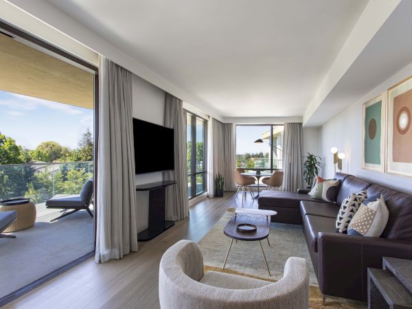 A modern living room with a balcony view, featuring a TV, sofas, armchair, abstract artwork, and a dining area with plants.
