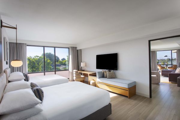 A modern hotel room with two beds, a TV, desk, and large window; connecting to a sitting area with more seating and windows.