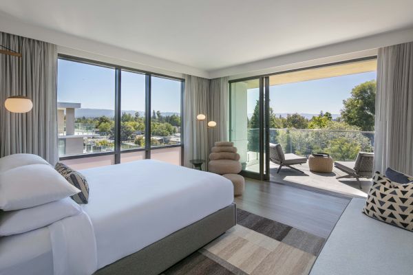 A modern bedroom with a large bed, panoramic windows, and a balcony with seating, overlooking a lush, green landscape.