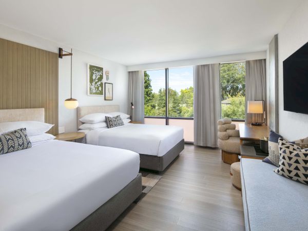 A modern hotel room with two beds, large windows, and daytime scenery. Comfortable seating and a TV are also present.