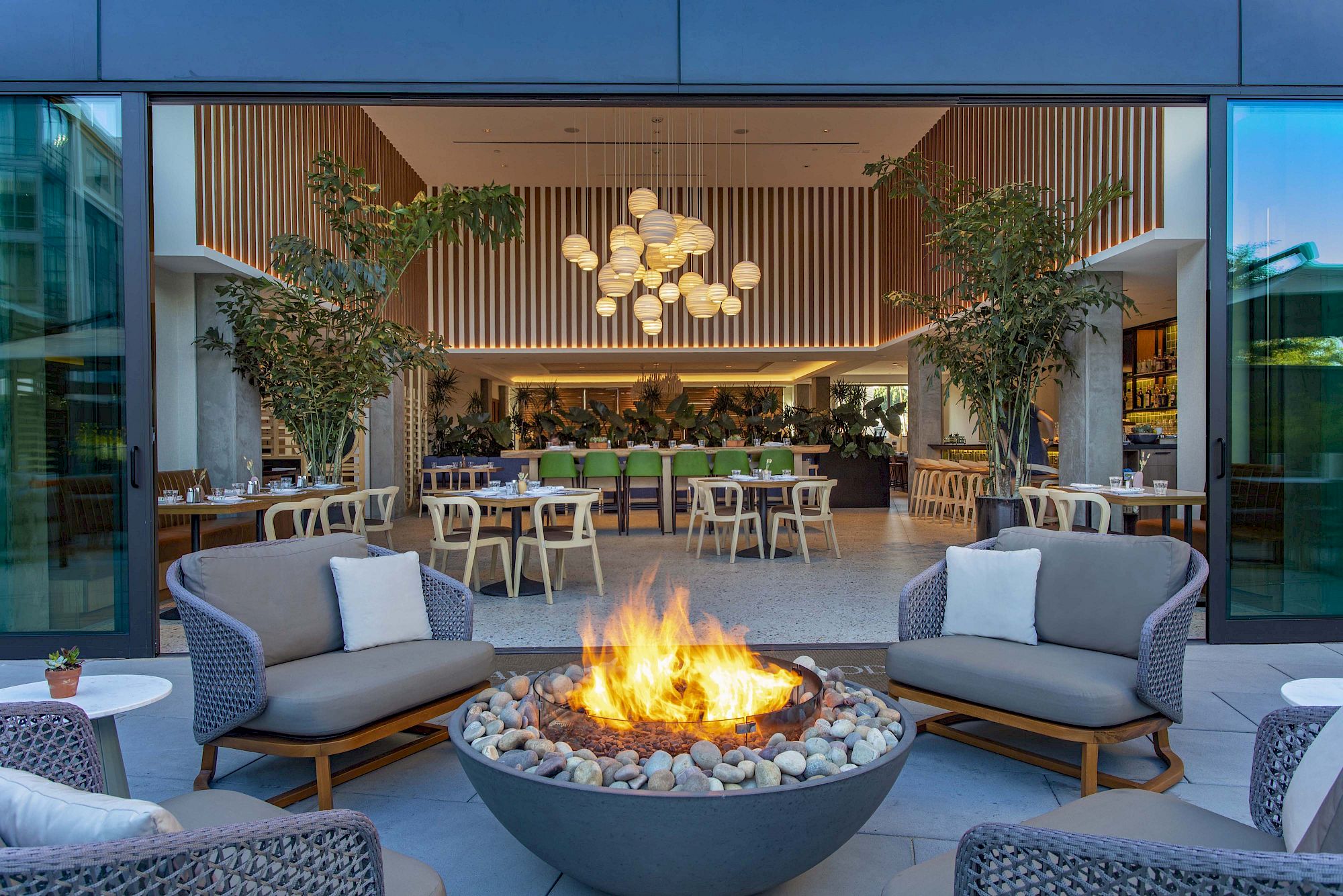 Outdoor seating with a fire pit and modern chairs, leading into an indoor restaurant with tables, plants, and hanging lights.