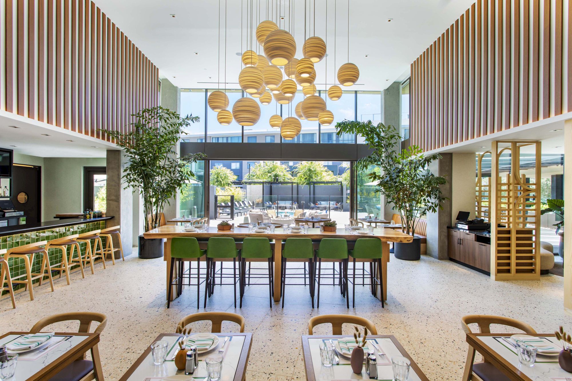 A modern restaurant interior with green chairs, wooden tables, hanging lights, and plants. Large windows show an outdoor area.