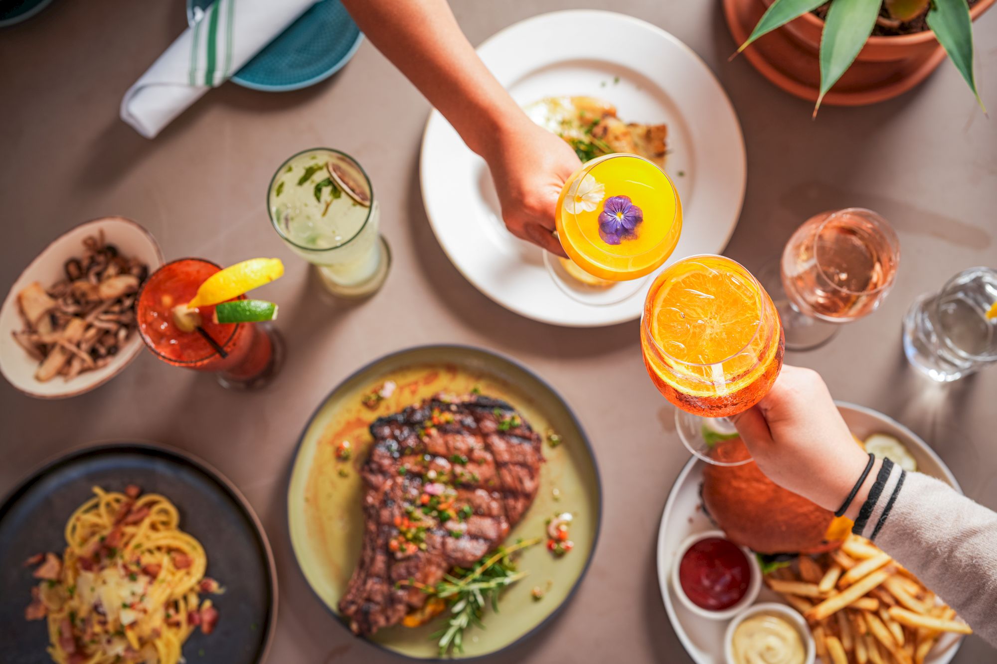 A group cheers with bright citrus drinks while plates of food cover the table in a lively meal moment.