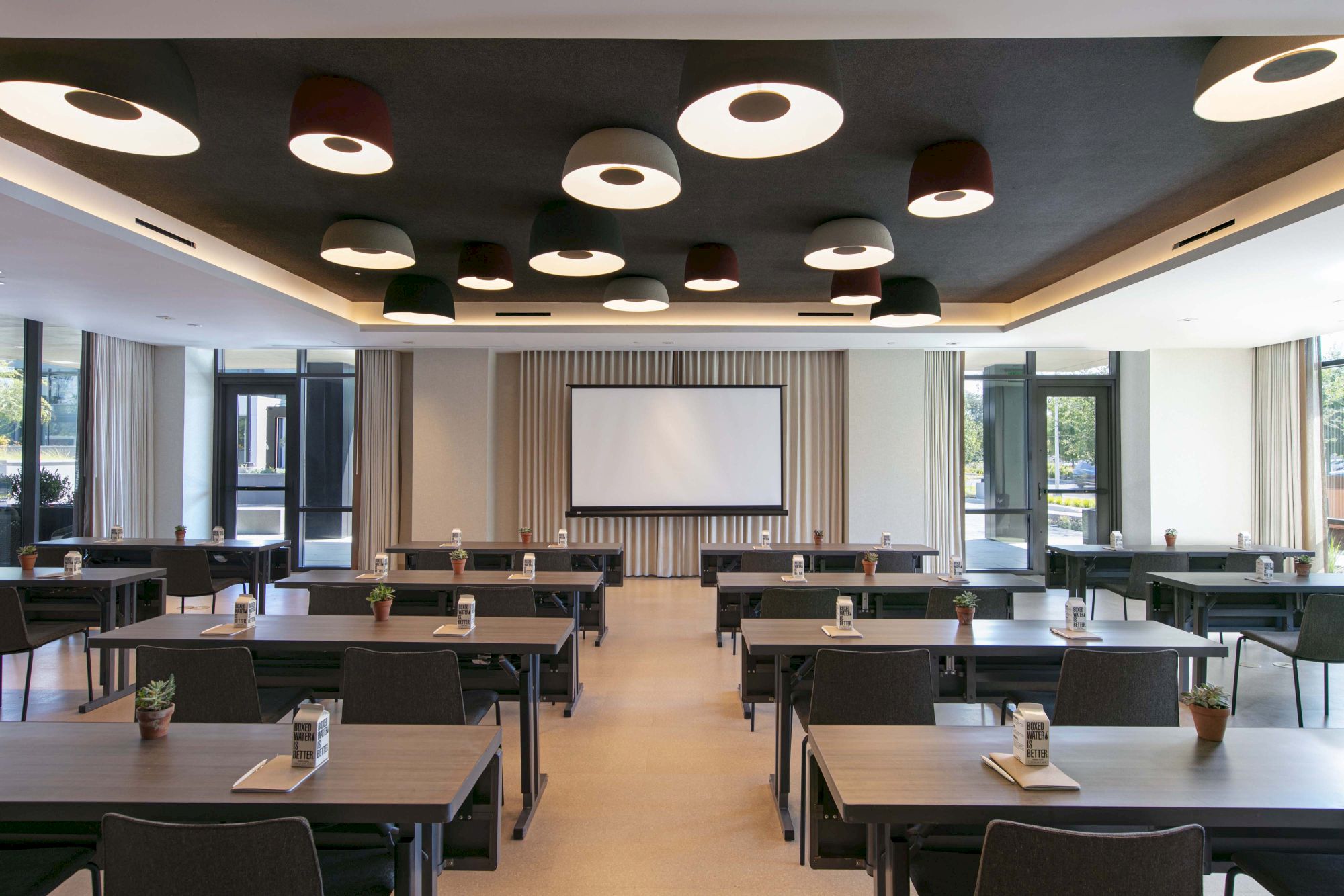 A modern conference room with tables, chairs, a projector screen, and potted plants on tables under stylish ceiling lights.