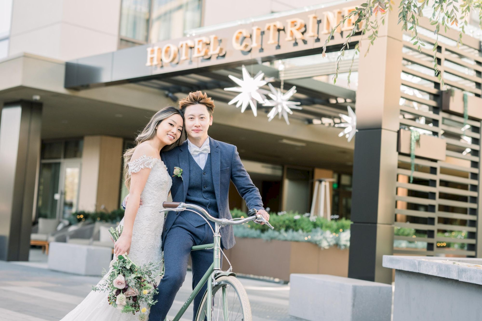 A couple in wedding attire is posing happily outside Hotel Citrine, with the groom on a bicycle and the bride holding a bouquet.