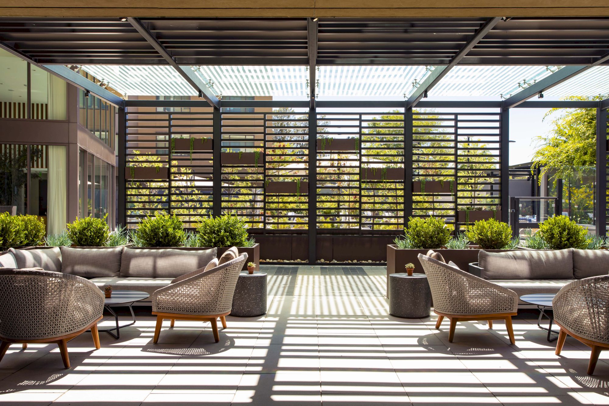 A modern outdoor patio features wicker furniture, potted plants, and a slatted roof, with sunlight casting shadows on the floor.