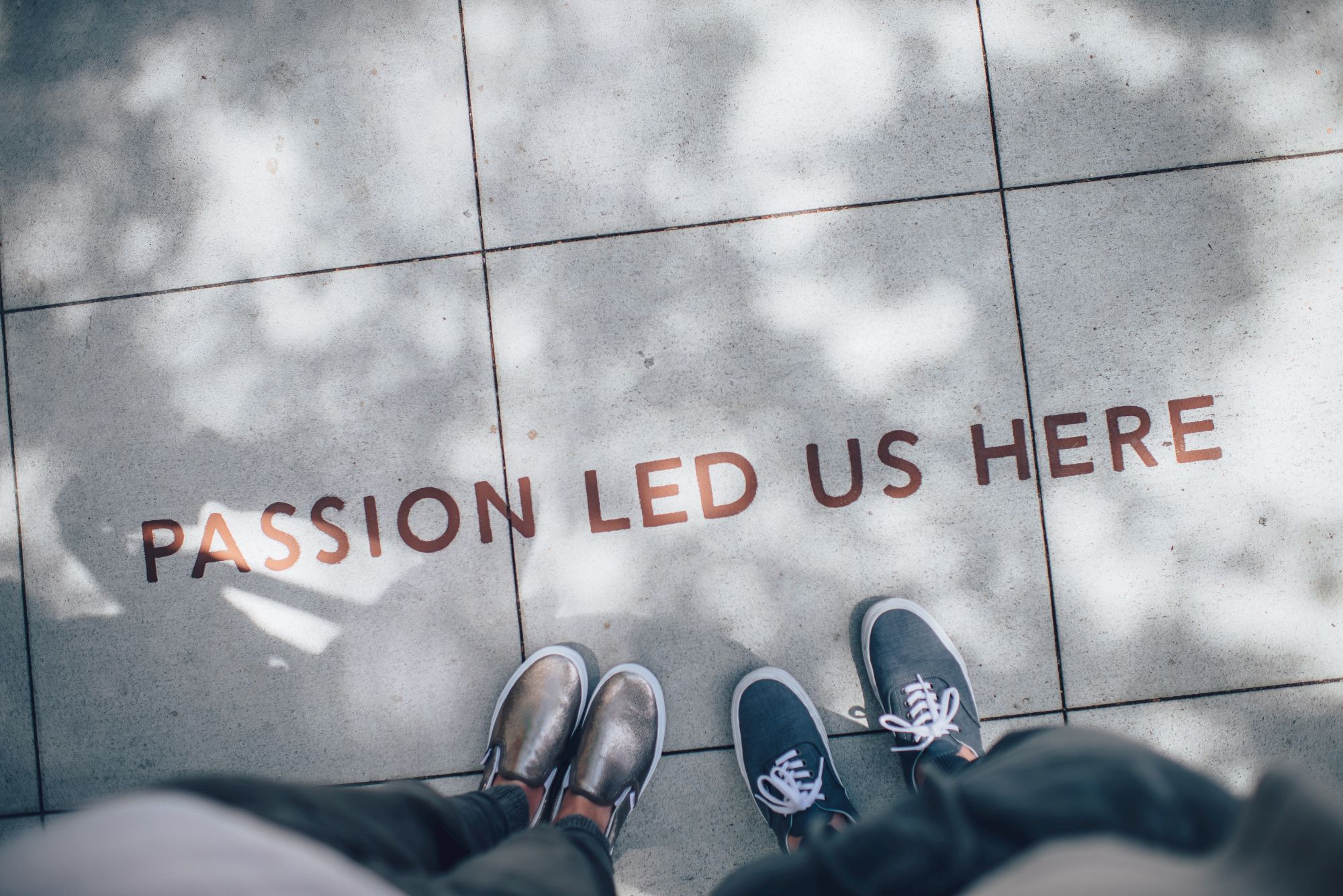 Two pairs of shoes stand on tiles with the words "PASSION LED US HERE" written on the ground.
