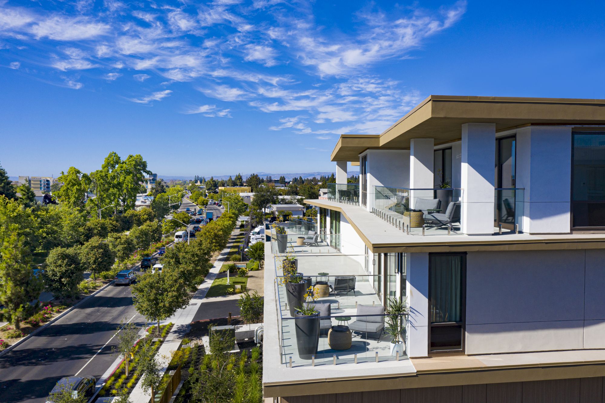A modern building&rsquo;s balcony overlooks a sunny city street lined with trees, cars, and distant buildings beneath a vast blue sky.