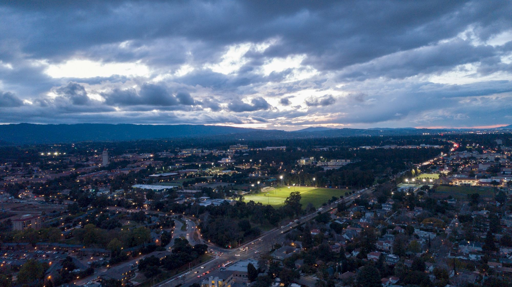 Aerial view of a cityscape at dusk, with illuminated fields, roads, and buildings under a cloudy sky.