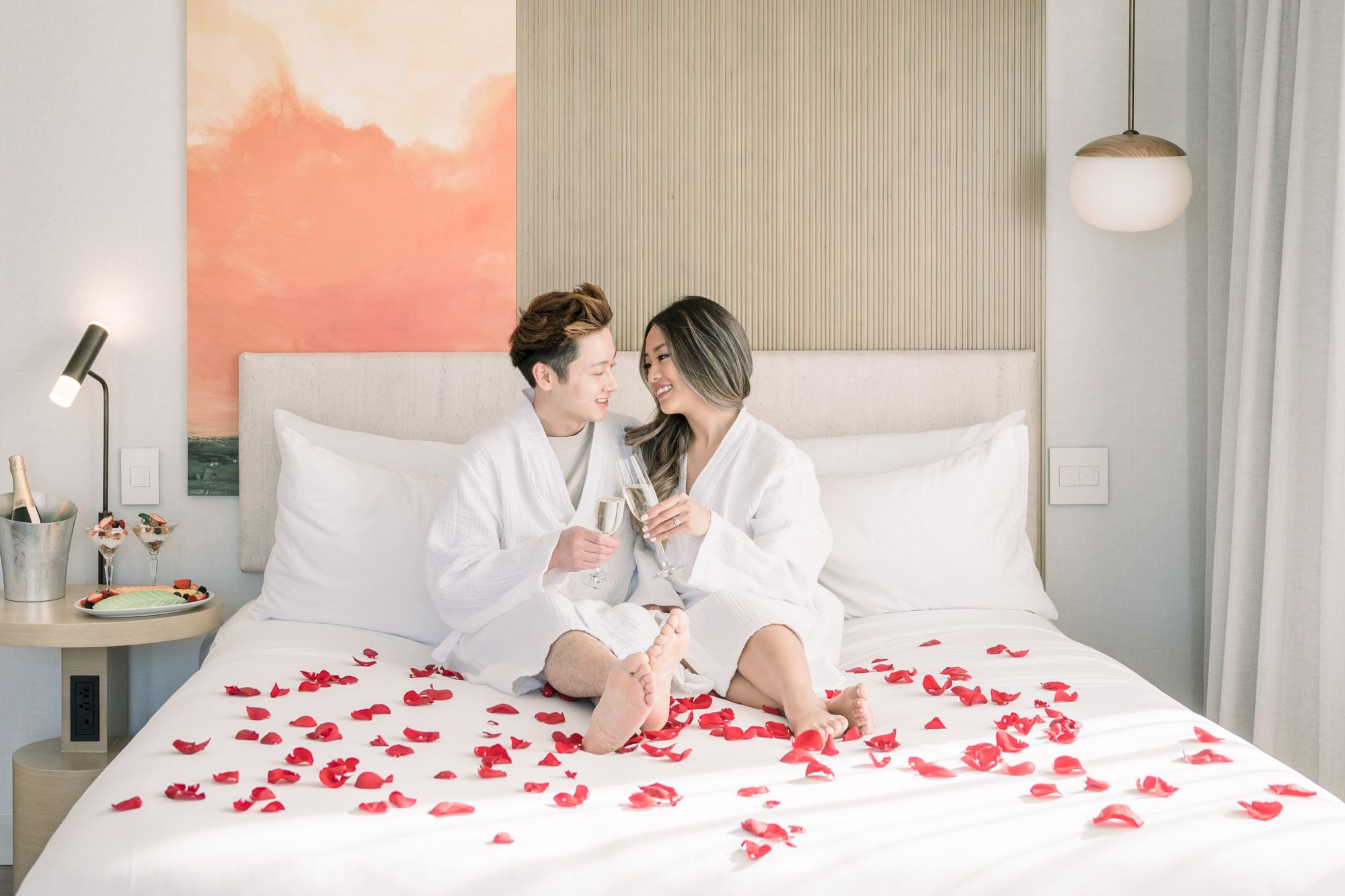 A couple in white robes sits on a bed adorned with red petals, toasting with glasses, under warm lighting in a cozy setting.