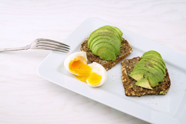 Sliced avocado on multigrain bread with a soft-boiled egg and a fork on a white plate.