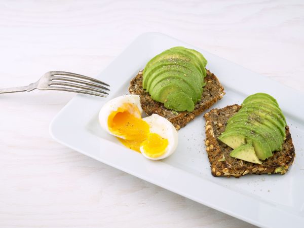 Sliced avocado on multigrain bread with a soft-boiled egg and a fork on a white plate.