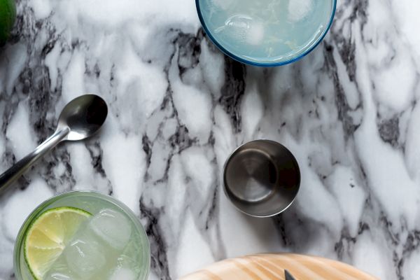 Two glasses with ice and lime on a marble surface, accompanied by a jigger, spoon, and cutting board with sliced limes and a knife.