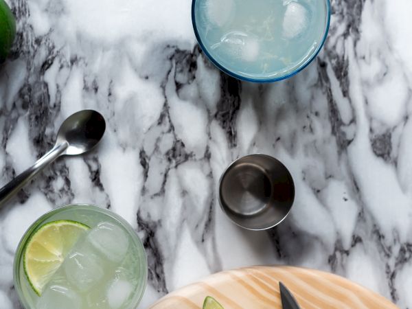 Two glasses with ice and lime on a marble surface, accompanied by a jigger, spoon, and cutting board with sliced limes and a knife.