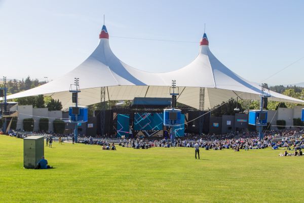 An outdoor venue with a large stage and canopy, surrounded by people seated on a grassy area, under a clear blue sky.