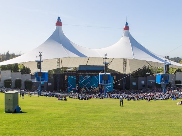 An outdoor venue with a large stage and canopy, surrounded by people seated on a grassy area, under a clear blue sky.