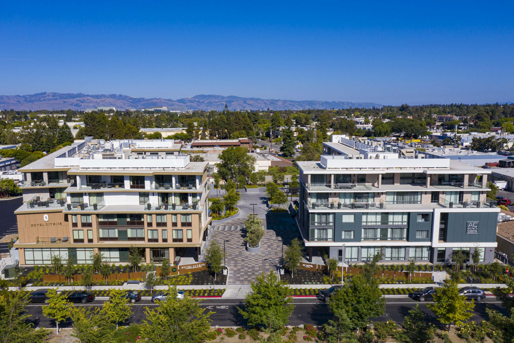 Two modern apartment buildings with balconies, trees lining the street, and mountains in the background under a clear blue sky.