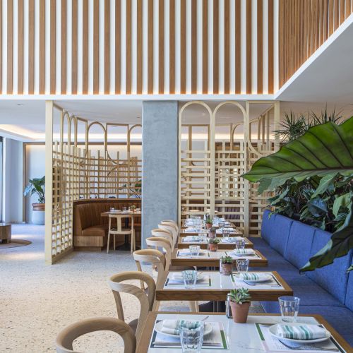 Modern restaurant with wooden decor, blue seating, and plants; tables set with plates, napkins, and glasses.