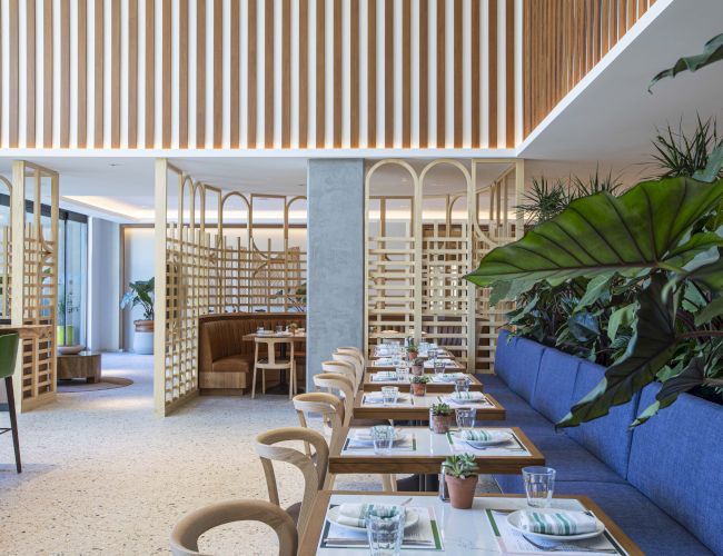 Modern restaurant with wooden decor, blue seating, and plants; tables set with plates, napkins, and glasses.