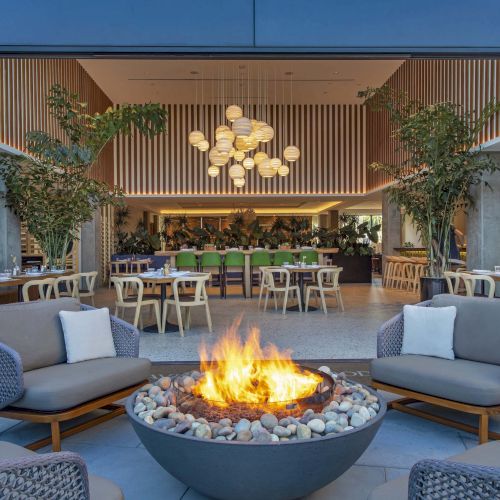 A stylish outdoor lounge with a fire pit, surrounded by sofas, leading into a modern indoor dining area with hanging lights and plants.