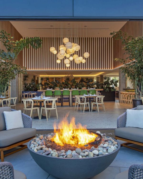A stylish outdoor lounge with a fire pit, surrounded by sofas, leading into a modern indoor dining area with hanging lights and plants.