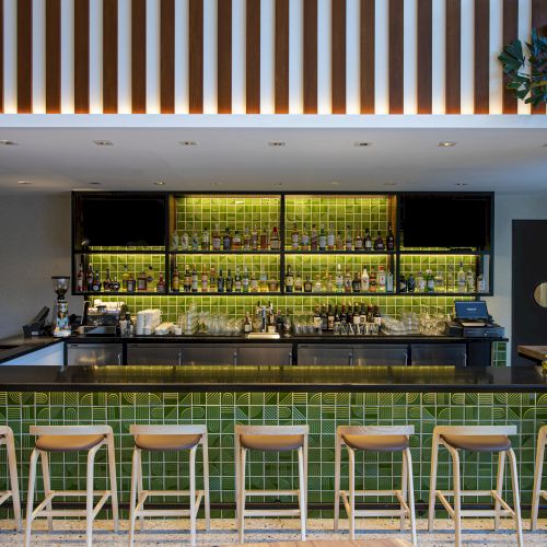 A modern bar with bright green backlit shelves of liquor bottles, a lit counter, ceiling lights, and wooden stools.