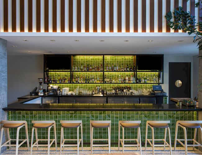 A modern bar with bright green backlit shelves of liquor bottles, a lit counter, ceiling lights, and wooden stools.
