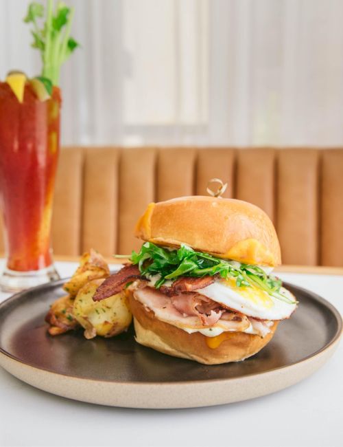 A burger with bacon, egg, and greens on a bun, served with potatoes on a plate, alongside a tall drink garnished with celery.