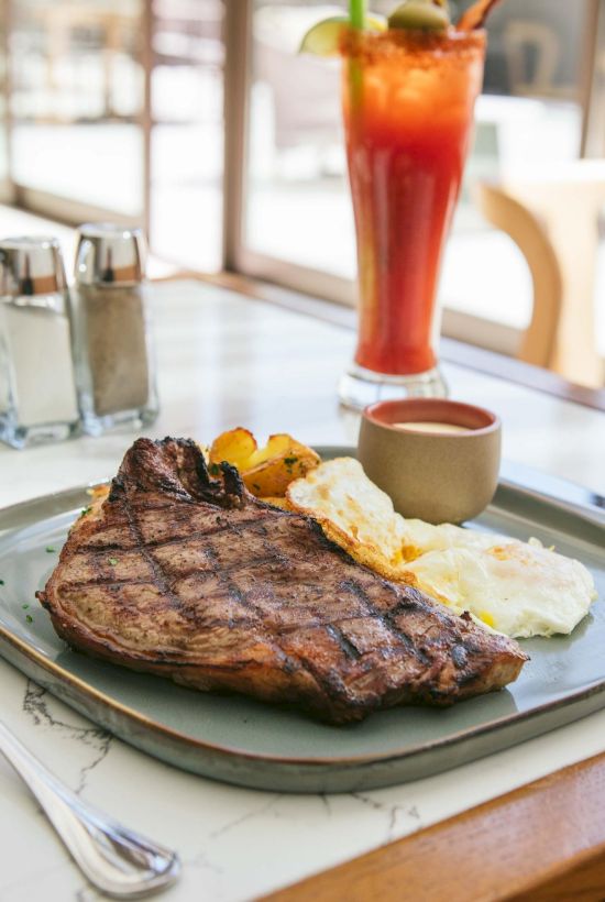 A plate with grilled steak, fried eggs, potatoes, and sauce, plus a tall red drink and salt and pepper shakers on a table.