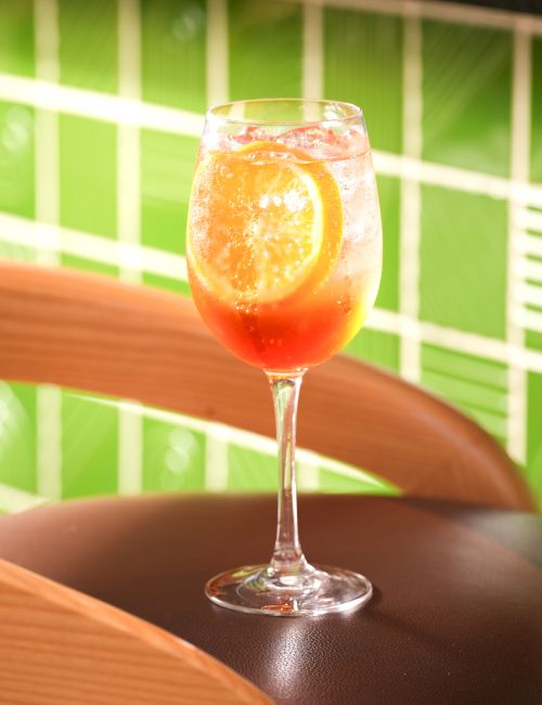 A stemmed glass filled with an orange-red iced drink sits on a wooden table, with a green-striped background behind it.