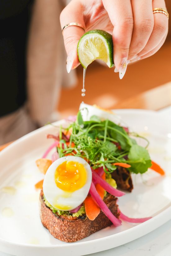 A hand squeezes a lime wedge, letting juice drip over a salad plate with greens, carrots, and a soft-boiled egg.