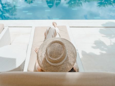 A person relaxes by a pool on a lounge chair, wearing a large straw hat, under a sunny sky with reflections of palm trees in the water.