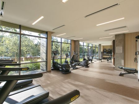 A bright, modern gym with large windows, treadmills, stationary bikes, and weight machines along a spacious, well-lit floor.