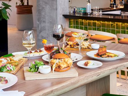 A modern dining setup with various dishes, including burgers and wine, on a wooden table surrounded by green chairs in a stylish interior.