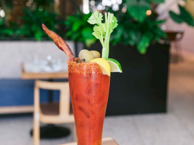 A Bloody Mary garnished with celery, olives, lemon, lime, and bacon, served in a tall glass on a table.