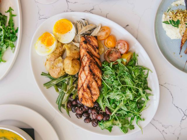 A colorful meal featuring grilled salmon, greens, olives, soft-boiled eggs, French fries, soup, wings, and a glass of rosé wine.
